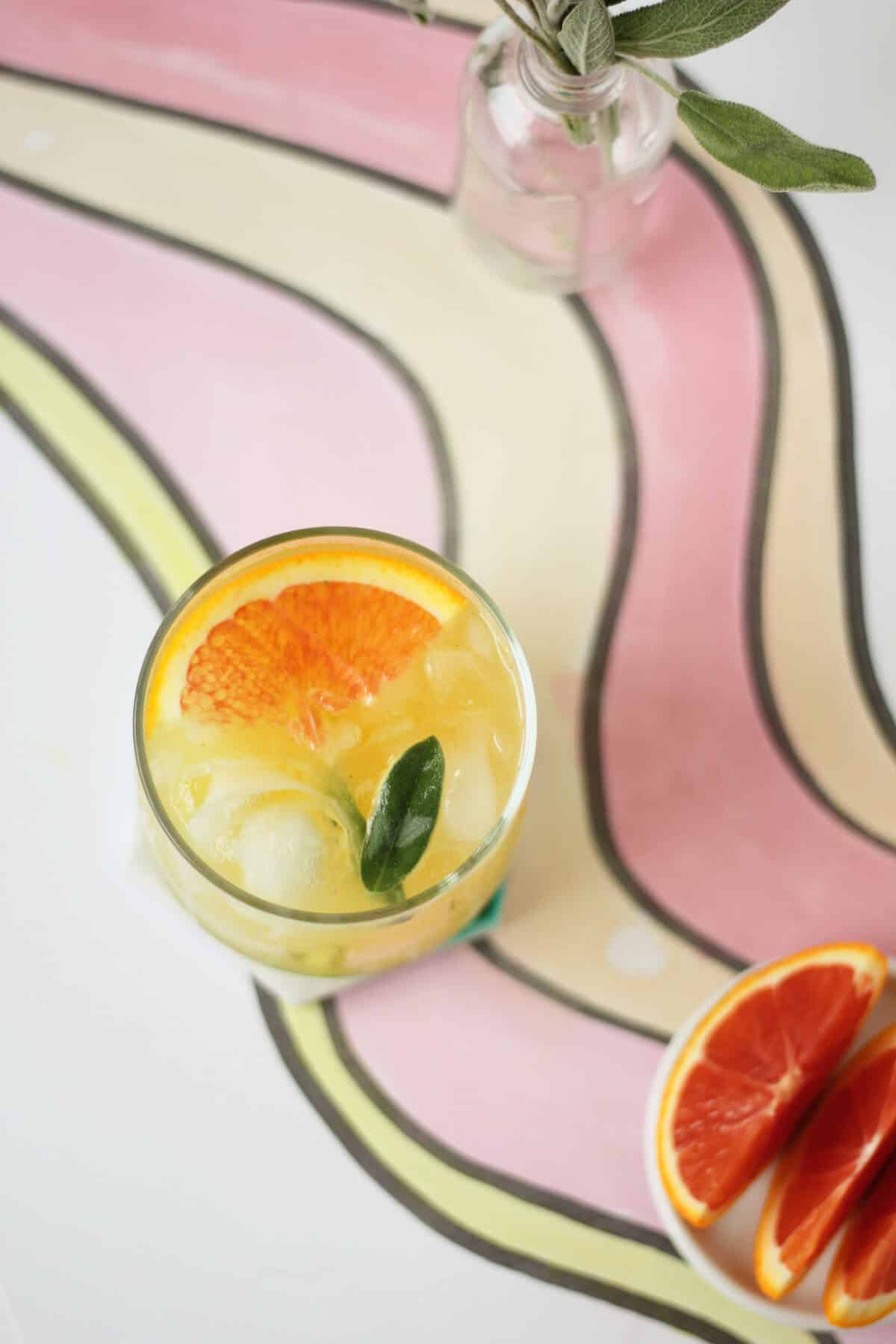 Overhead shot of a clear glass cup filled with ice, orange liquid, an orange slice, and a sage leaf. There's a mini bottle with a sprig of sage stuck in it, as well a dish with a few wedges of blood orange on it.