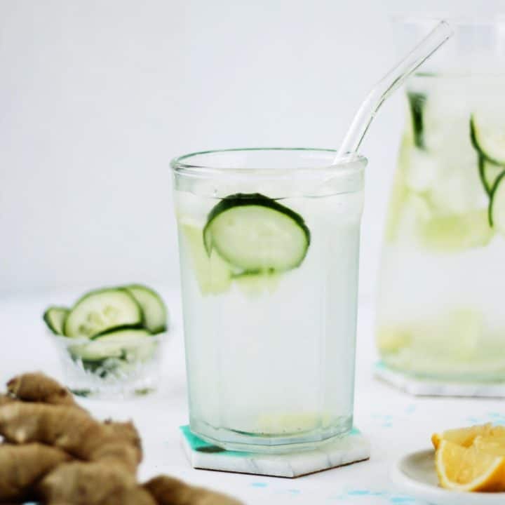 Clear glass cup filled with water and a slice of cucumber, with a glass straw. There is some fresh ginger root and a dish of lemon wedges in the foreground, with a cup of cucumber slices and a pitcher of water in the background.