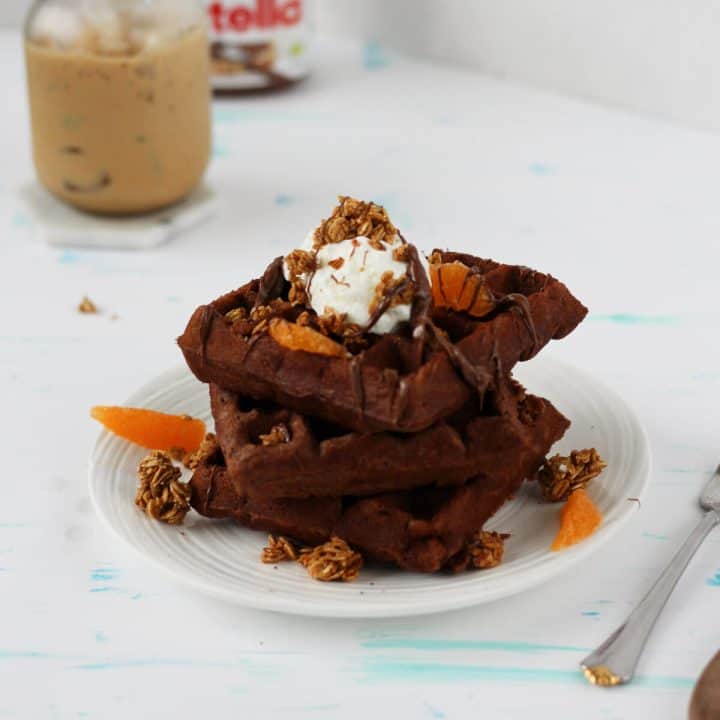 Stack of brown waffles with Nutella dripping off them and topped with a scoop of yogurt, orange slices and granola. There's a cup of ice coffee and a jar of Nutella in the background.