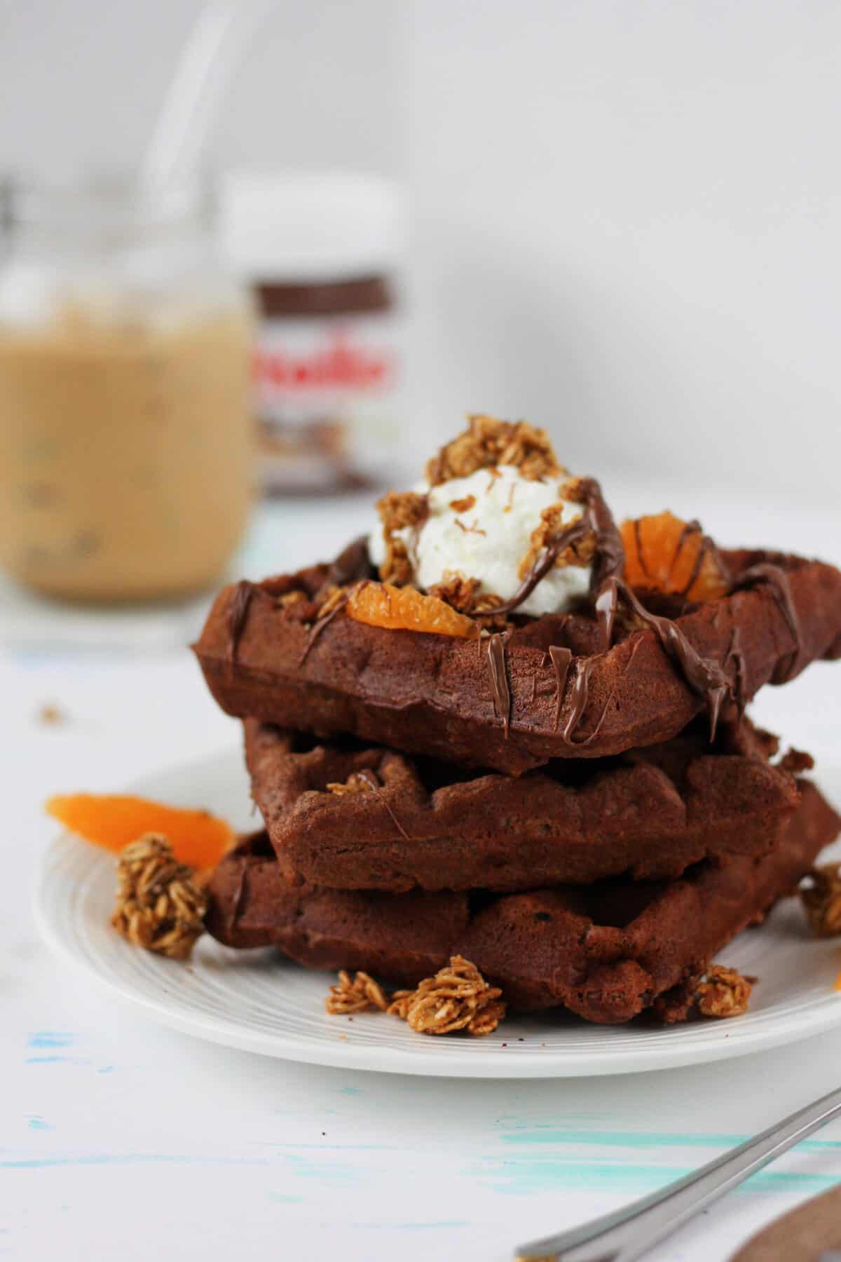 Stack of brown waffles with Nutella dripping off them and topped with a scoop of yogurt, orange slices and granola. There's a cup of ice coffee and a jar of Nutella in the background.