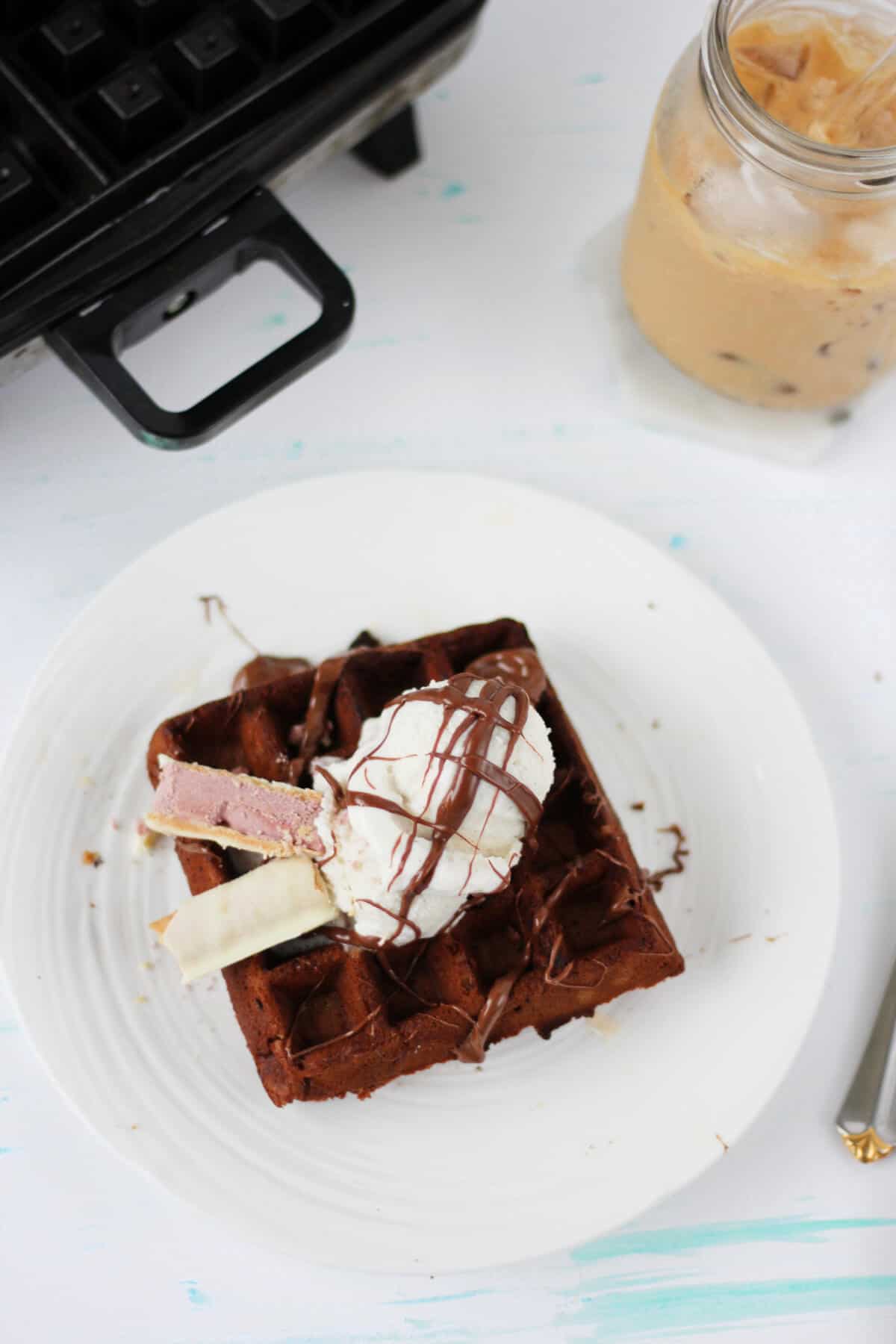 Overhead view of one waffle topped with vanilla ice cream, Nutella and a cigar cookie. A waffle maker and a glass of iced coffee are on the edges of the picture.