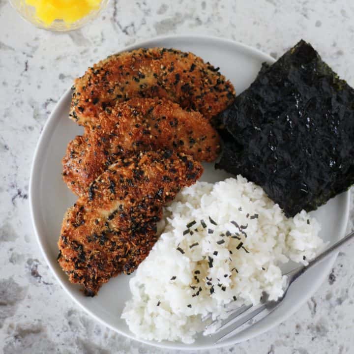 White plate with three big furikake chicken tenders, rice sprinkled with sesame seeds and a few small sheets of seaweed. There's a bowl of marinated bean sprouts, as well as a smaller bowl of bright yellow pickled radish, in the background.